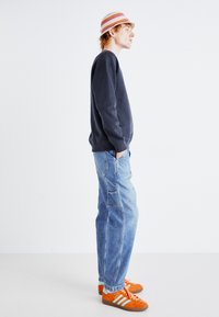 Navy sweatshirt, light blue baggy jeans, orange sneakers with blue stripes, and a multicolored striped bucket hat. Side profile view.
