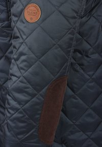 Marineblå quiltet jakke med en brun fløjlsdetalje på lommen og en rund læderbrandpatch. Glat tekstur og diamantmønster.