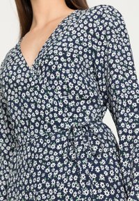 Vestido azul-marinho com estampa floral branca, decote em V, mangas longas e detalhe de cintura atada. O tecido é leve com uma textura suave.