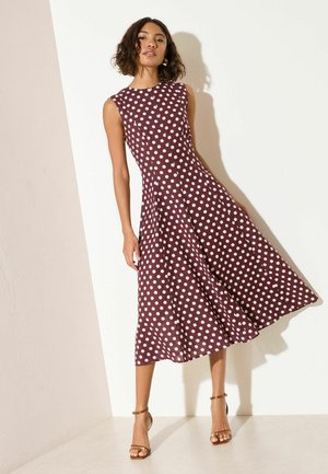 Kobieta w bordowej sukience midi bez rękawów w białe kropki, pozująca na tle białej ściany, w połączeniu z beżowymi szpilkami na pasku wokół kostki.