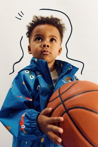 Veste imperméable bleue avec des motifs colorés sur le thème du sport, des poignets élastiques et une fermeture éclair à l'avant, tenue par un enfant avec un ballon de basket.