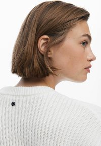 Witte ribgebreide trui met een ronde hals en een klein zwart logo accent aan de achterkant. Zichtbaar getextureerd breipatroon en cropped ontwerp.