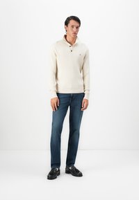 Homme portant un pull crème côtelé avec col et boutons, un jean bleu et des mocassins à pampilles noirs, se tenant contre un fond blanc.