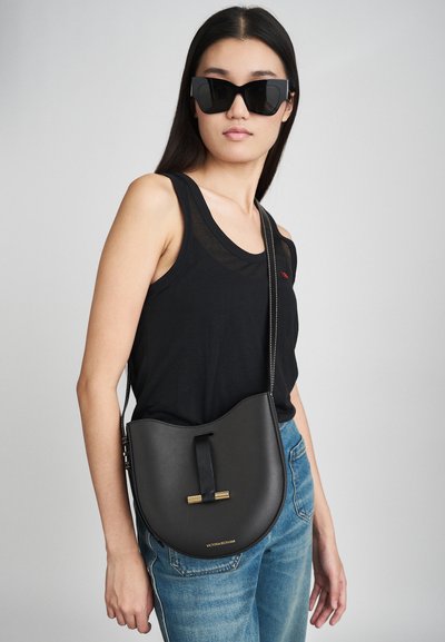 Melnas ādas sēžamais maisiņš ar izliekta silueta, zelta aksesuāriem un noņemamu plecu siksnu. Valkā ar melnu topi un ziliem džinsiem.