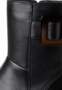 Bottine en cuir noir avec une texture lisse, présentant un détail de boucle rectangulaire sur le côté et des coutures contrastantes.