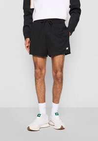 Shorts de sport en mesh noir avec taille élastique et cordon de serrage. Dotés d'un logo sur la jambe gauche, ils sont assortis de chaussures et de chaussettes blanches.