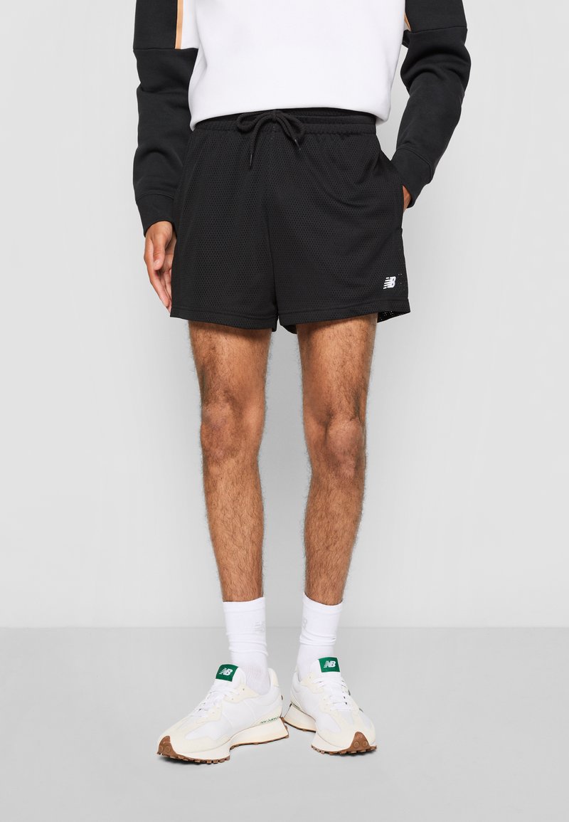 Shorts de sport en mesh noir avec taille élastique et cordon de serrage. Dotés d'un logo sur la jambe gauche, ils sont assortis de chaussures et de chaussettes blanches.