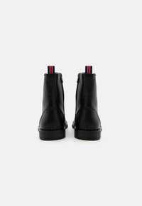 Botas de tobillo de cuero negro con una textura suave, cierre de cremallera y una tira de tela de tirador con rayas rojas y blancas en la parte trasera.