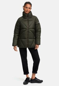 Dunkelgrüne Pufferjacke mit hohem Kragen, diagonalem Steppmuster, Reißverschluss und zwei Fronttaschen. Kombiniert mit gerollten schwarzen Hosen und Schuhen.