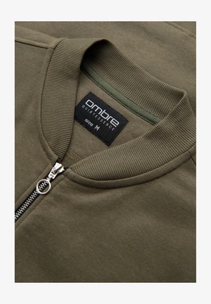 Oliwkowa kurtka bomberka z prążkowanym kołnierzem i zamkiem. Metka wskazuje markę "Ombre" i rozmiar "M." Gładka faktura materiału.