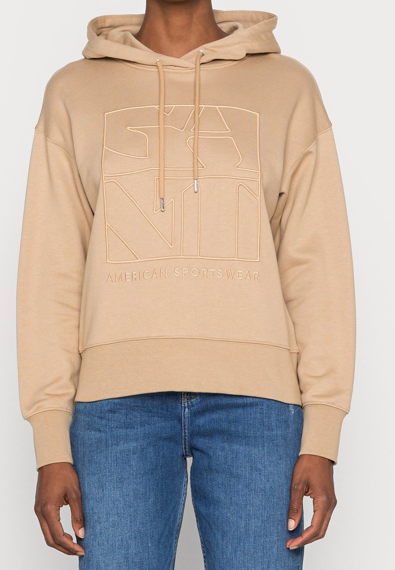 Sweat à capuche beige avec un grand logo en relief "GANT" à l'avant et le texte "American Sportswear", porté avec un jean bleu par une personne debout.