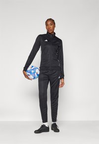 Survêtement de sport noir avec des rayures blanches, comprenant une veste à fermeture éclair et un pantalon fuselé. La personne tient un ballon de football bleu et rouge.