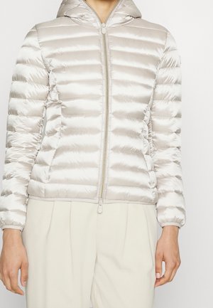 Femme portant une veste matelassée à capuche beige clair avec une fermeture éclair complète, associée à un pantalon plissé de couleur claire.