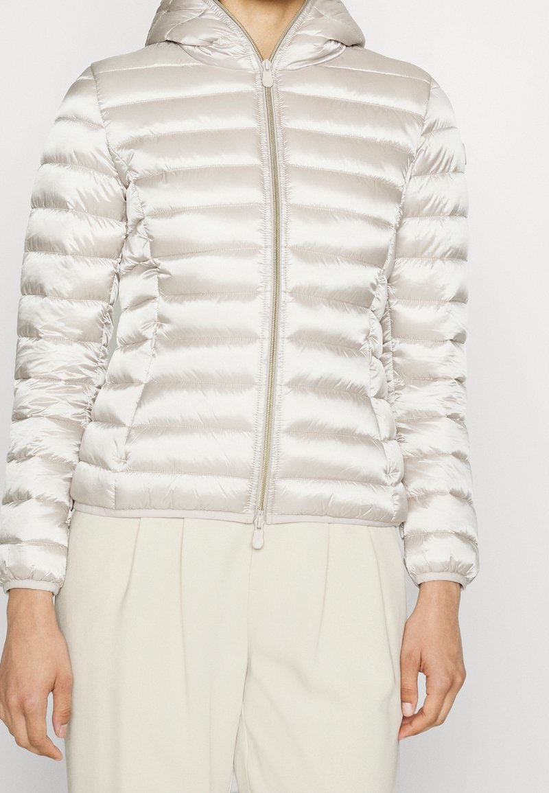 Femme portant une veste matelassée à capuche beige clair avec une fermeture éclair complète, associée à un pantalon plissé de couleur claire.