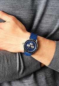 Blaues Gummiband-Uhr mit einem runden, dunkelblauen Zifferblatt, silberfarbenem Gehäuse und drei Unterzifferblättern. Am Handgelenk über einem grauen Pullover getragen.