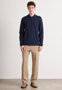 Polo de manga longa azul-marinho com colarinho com fecho, tecido texturizado. Combinado com calças bege e sapatos de camurça castanhos, em pé contra um fundo neutro.