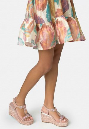 Piernas con sandalias de cuña nude con tiras adornadas con joyas, combinadas con un vestido floral hasta la rodilla con dobladillo con volantes sobre un fondo liso.