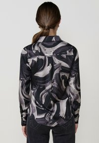 Camicetta a maniche lunghe con un motivo astratto in bianco, grigio e nero. Realizzata in tessuto liscio con colletto e polsini classici.