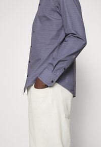 Polohemd in paars met een geometrisch patroon, lange mouwen en zwarte knopen. Gepaard met lichtgekleurde broeken.