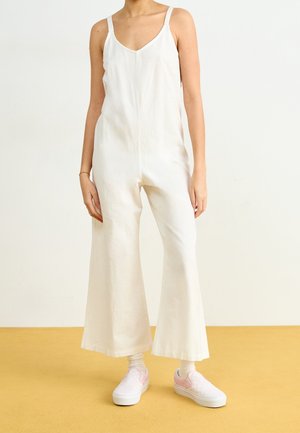 Biały kombinezon lniany z szerokimi nogawkami, cienkimi ramiączkami i dekoltem w kształcie litery V, w połączeniu z jasnoróżowymi butami wsuwanymi. Gładka faktura, bez widocznych wzorów.