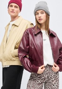 Νεαρός άνδρας με μπεζ σακάκι και κόκκινο σκουφάκι στέκεται δίπλα σε γυναίκα με σκούρο κόκκινο δερμάτινο μπουφάν, γκρι σκουφάκι, λευκό πουκάμισο και παντελόνι με λεοπάρ σχέδιο.