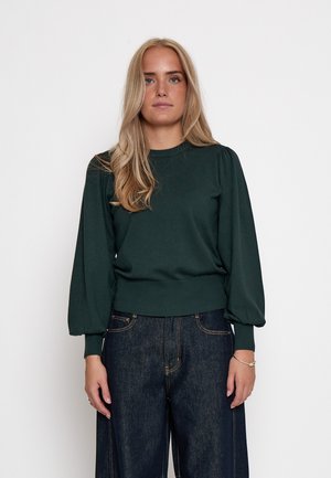 Mujer rubia con un suéter de manga larga verde oscuro y jeans oscuros de pierna ancha, de pie frente a un fondo blanco liso.