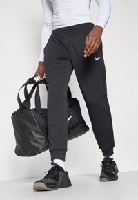 Μαύροι αθλητικοί παντελόνες Nike με ελαστική ζώνη, κωνικές γάμπες και πλαϊνές τσέπες. Σκούρος μαύρος σάκος Nike κρατημένος με το ένα χέρι; μαύρα αθλητικά παπούτσια με λεπτομέρειες.