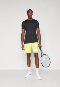 Chemise noire à manches courtes et short jaune fluo avec le texte "BOR". Baskets blanches et une raquette tenue dans la main droite, se tenant contre un fond gris.