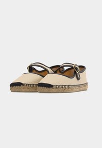 Espadrilles plates en toile beige avec bouts noirs, bordures noires, brides à la cheville, boucles dorées et semelles en jute tressé sur fond blanc.