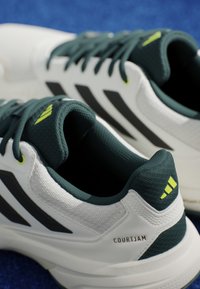 Baskets de tennis blanches et vertes foncées avec une tige en maille texturée, des rayures noires et un accent jaune sur la languette. Présente le logo "COURTJAM".