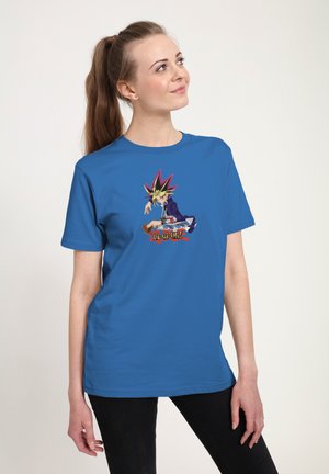 Kvinde med langt hår bundet tilbage iført en blå T-shirt med et farverigt grafisk motiv af en Yu-Gi-Oh karakter og sorte bukser foran en ensfarvet baggrund.