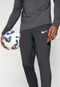 Sorte træningsbukser med et hvidt Nike-logo og en tætsiddende design, parret med en grå langærmet skjorte, idet man holder en mønstret fodbold.