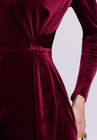 Burgundowa aksamitna sukienka z fantazyjnym węzłem w talii i długimi rękawami o gładkiej, błyszczącej fakturze.