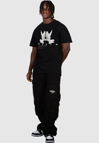 T-shirt noir avec impression graphique blanche de deux oiseaux et "YOUTH", associé à un pantalon cargo noir avec des poches latérales et un détail brodé.