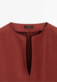 Détail du col d'une chemise en lin couleur rouille avec un col rond et une fente en V à l'avant, montrant une étiquette noire à l'intérieur de l'encolure.