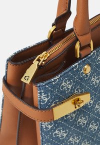 Torba z dżinsu z jasnobrązowymi skórzanymi wstawkami, wyposażona w złote okucia, zapięcie na suwak oraz misterny biały kwiatowy wzór.