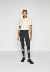 Lichtbeige cropped t-shirt met korte mouwen, gecombineerd met donkergrijze leggings met lichtgrijze paneelaccenten. Model draagt zwarte schoenen.