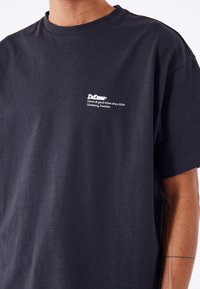 T-shirt in cotone nero a maniche corte, con un logo stampato bianco sul lato sinistro del petto. Texture liscia, vestibilità morbida.