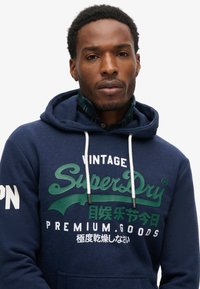 Felpa con cappuccio blu navy con dettagli del logo in verde e bianco. Presenta un cappuccio regolabile e un tessuto morbido e testurizzato. Vestibilità atletica classica.