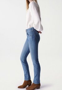 Světle modré džínové skinny džíny s vysokým pasem, s klasickým 5-kapsovým designem, v kombinaci s bílou blůzou a hnědými semišovými kotníkovými botami.
