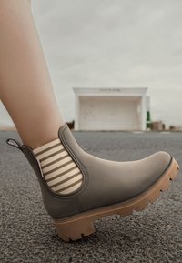 Botas de tobillo impermeables grises con un upper de goma texturizada, forro de tela a rayas y una suela de plataforma marrón, que incluye una lengüeta trasera.