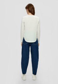 Haut à manches longues blanc avec un ourlet arrondi, associé à un jean bleu foncé ample et des baskets blanches ; tissu lisse et design décontracté.