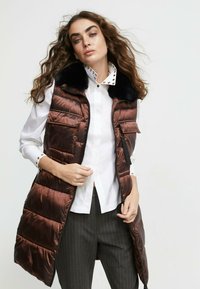 Brown puffer vest with a shiny finish, a black fur collar, and front pockets. Worn over a white shirt with studded collar and dark striped pants.