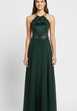 Jeune femme portant une robe de soirée sans manches vert foncé avec un corsage en dentelle et un décolleté à fines bretelles, debout et souriante sur un fond uni.
