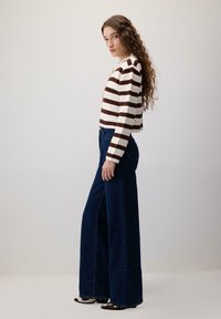 Pulover tricotat cu dungi în crem și maro, croială scurtă; blugi evazați, de culoare albastru închis, cu talie înaltă și slituri laterale; pantofi plate cu accente texturate.