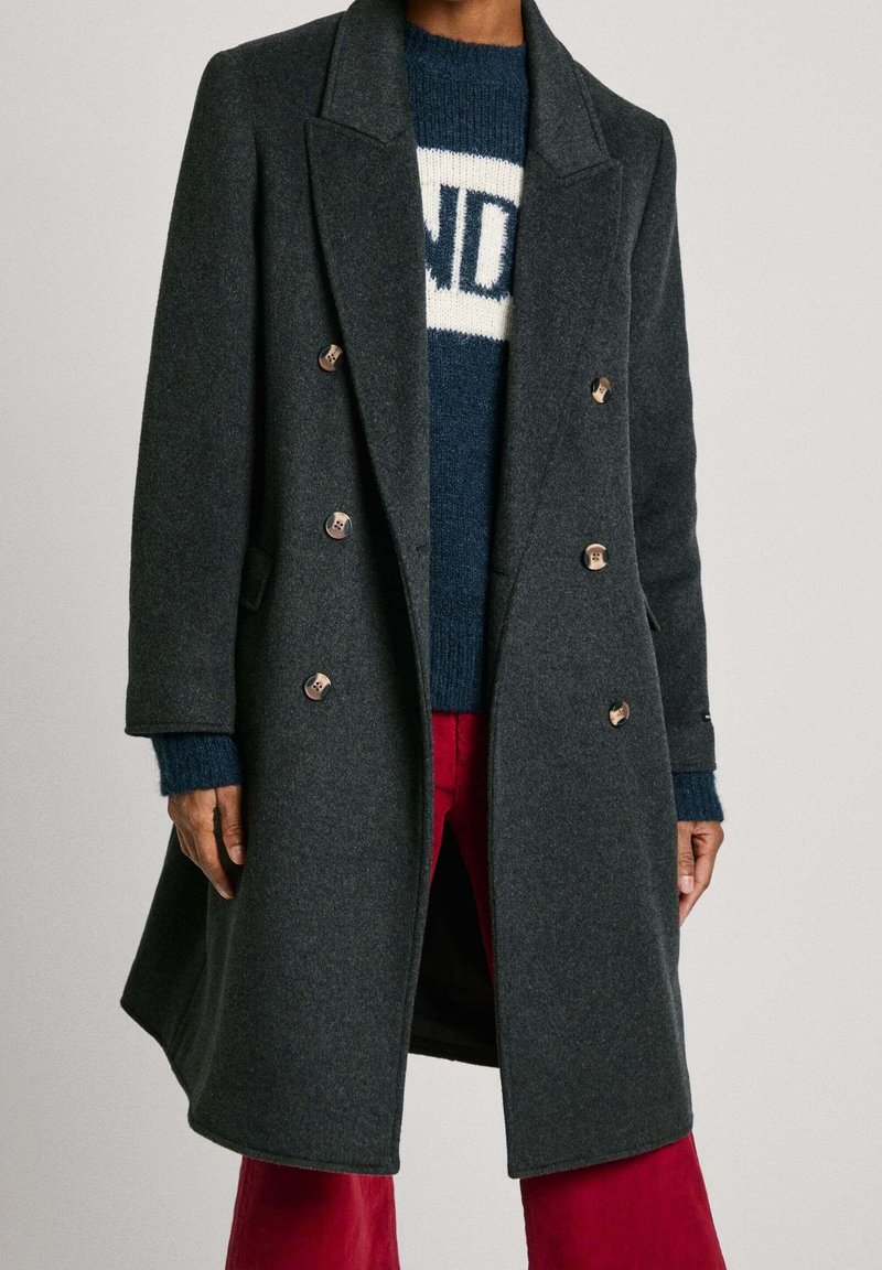 Grijze wollen jas met een dubbelgeknoopte sluiting, een doorgestikte kraag en metalen knopen. Draagt over een blauwe trui en rode broek.