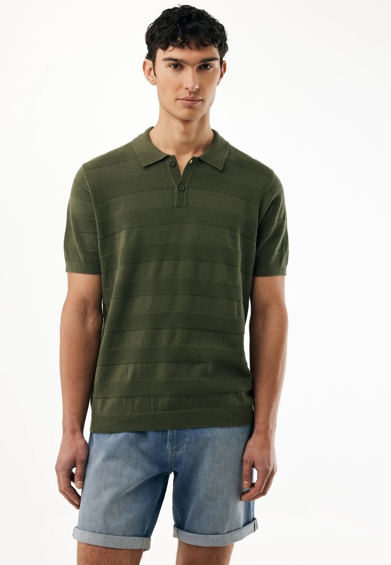 Giovane uomo che indossa una polo verde a maniche corte e pantaloncini di jeans azzurri arrotolati, in piedi contro uno sfondo bianco.