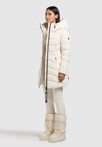 Puffer bunda v svetlo bielej farbe s prešívaným vzorom, vysokým golierom, predným zipsom a bočnými vreckami. Kombinovaná s ponožkovými snehovými čižmami po kolená vo farbe bundy.