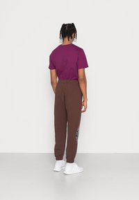 T-shirt de algodón morado combinado con pantalones de chándal marrones que tienen un gráfico colorido en la pierna izquierda y una cintura elástica. Zapatillas blancas.