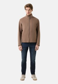 Bruine jas met rits en een hoge kraag, gemaakt van zacht materiaal; gecombineerd met donkere spijkerbroek en lichtgekleurde schoenen. Geen zichtbare patronen of accenten.